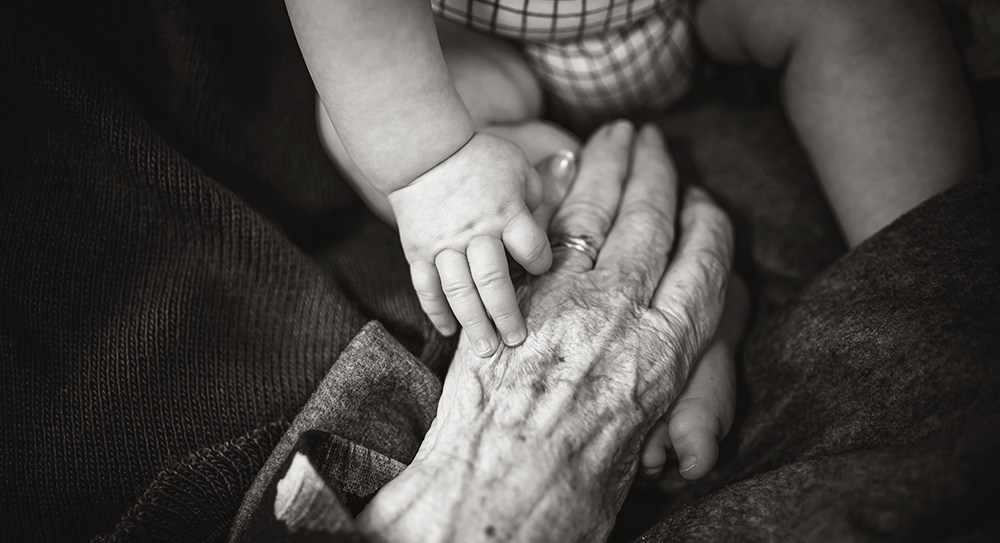 A black and white image of an baby's hand gripping the little finger of a more aged hand.