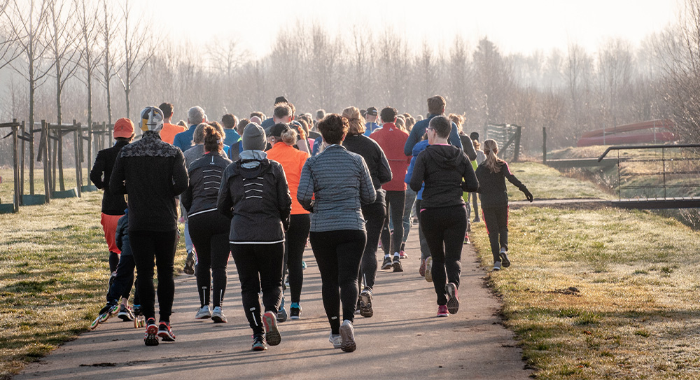 Image taken of the back of a large group of runners running along a wide path in a park. There are trees in the distance and a hazy light with lots of mist - it looks cold and frosty on the grass either side of the path. 