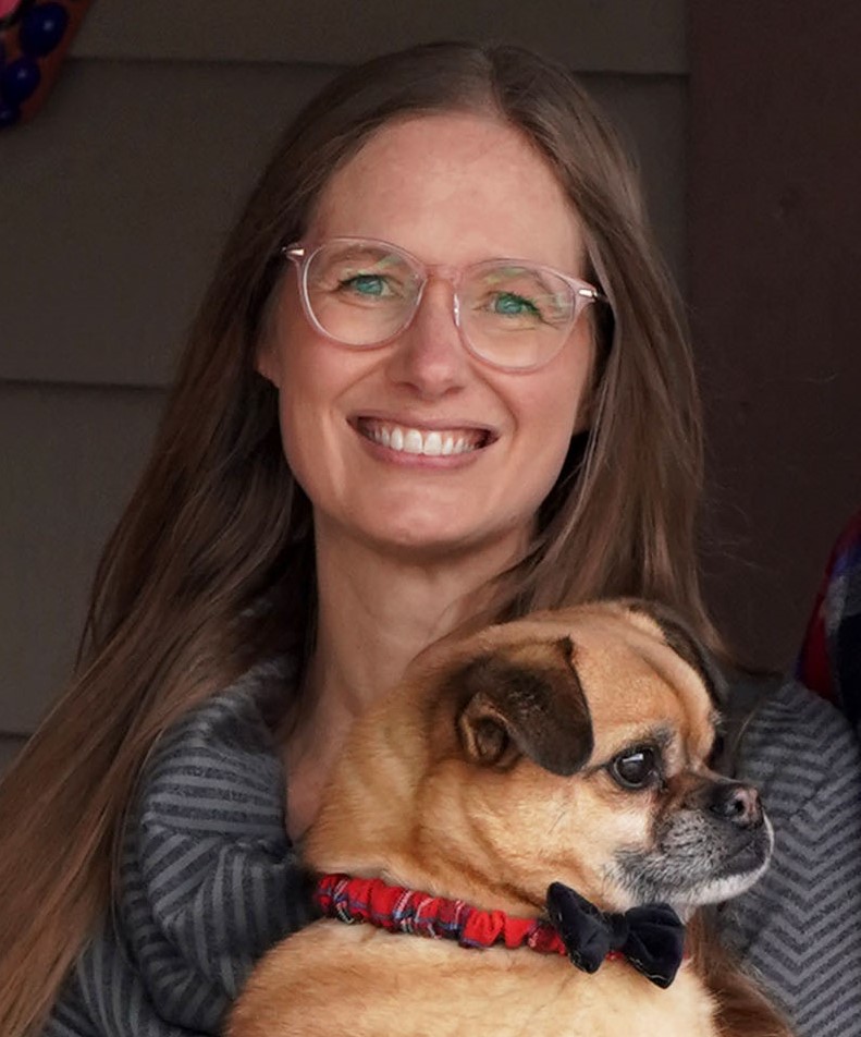 The image features a woman wearing glasses smiling while standing next to a dog. The woman is indoors with a wall in the background. The dog is wearing a red collar.