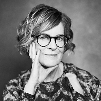 The image shows a woman wearing glasses indoors. She is smiling and the photo is in black and white.