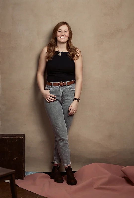 The image features a woman standing in front of a wall indoors. The person is wearing jeans and casual clothing, with a smile on their face.