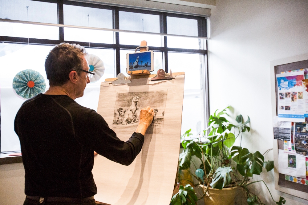 The image features a man drawing with charcoal on a sheet of paper attached to an easel. The setting includes indoor elements such as a houseplant, flowerpot, vase, art, and a window.