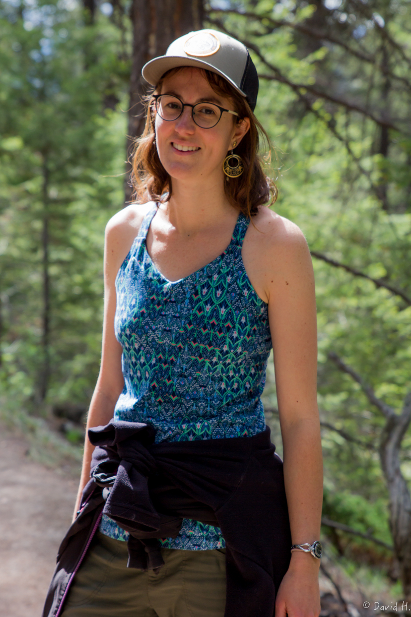 The image features a woman wearing a hat and glasses while standing in a forest. She is smiling and appears to be enjoying the outdoors.