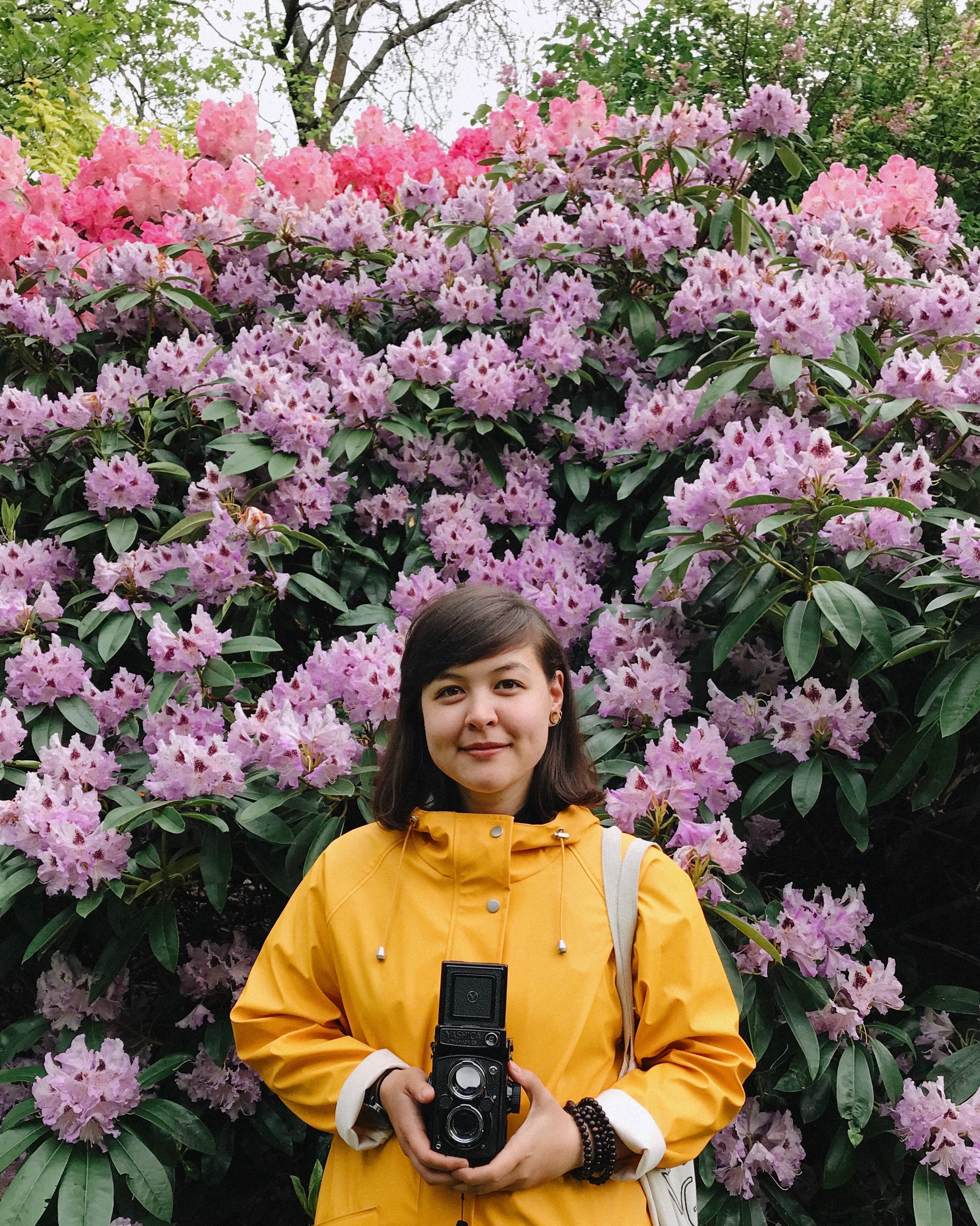 The image shows a person holding a camera outdoors. She is wearing a yellow jacket and the background includes a tree and flowers. The colors pink and purple are present in the image, suggesting a springtime setting.