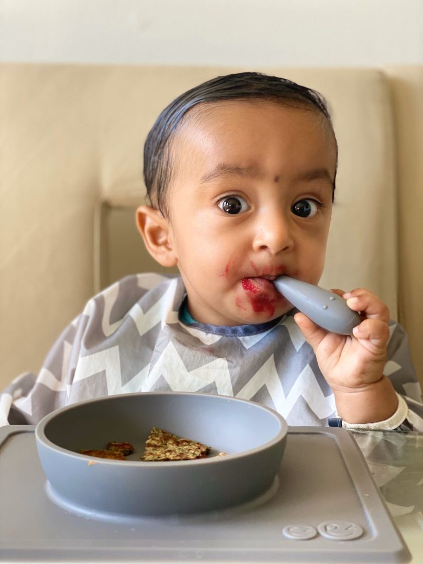 Photograph of a baby feeding himself using the ezpz Tiny Spoon with food placed in the ezpz Mini Bowl. The food pictured is a traditional Newari dish.