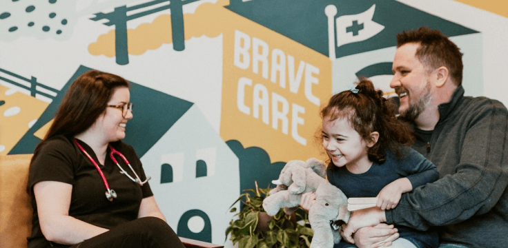 Photo of a father holding a child talking to a Brave Care provider in front of a mural