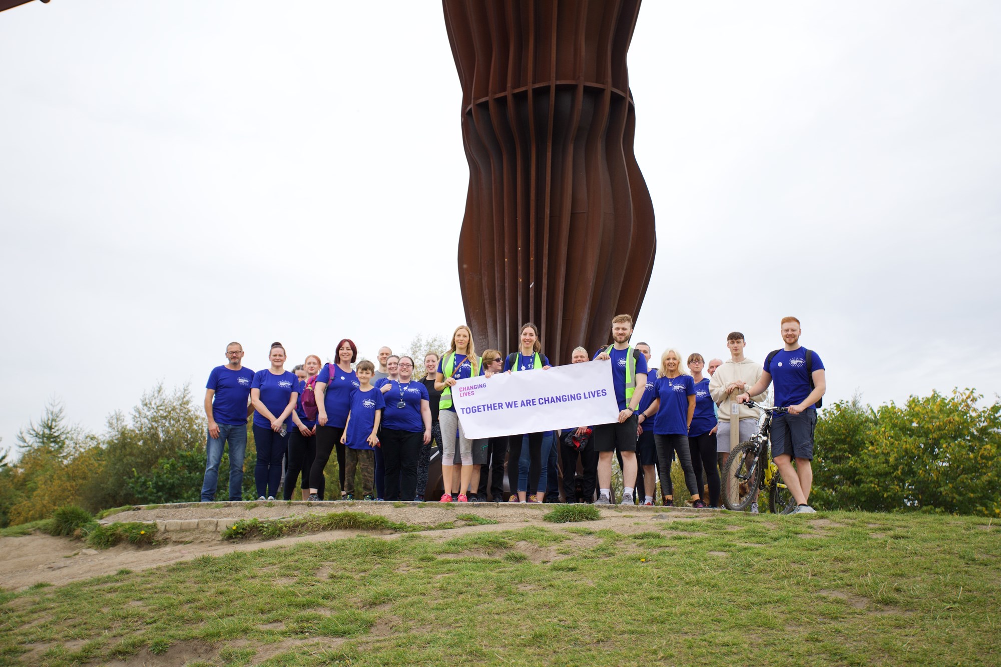 Changing Lives staff members stood in front of the Angel of the North after completing the staff walk of their annual fundraiser, 50K Your Way, holding a banner which reads "Together We Are Changing Lives."