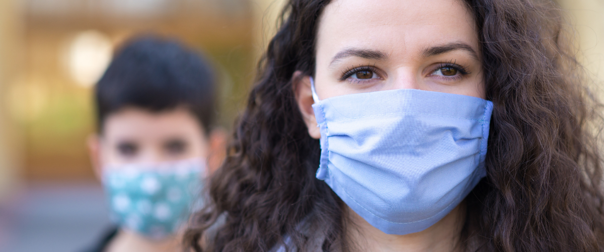 A woman in the foreground wearing a blue mask and another person in the background, blurred, also wearing a blue mask