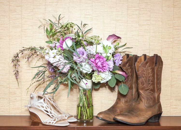 Açık renk arka plan önünde, bir vazo içerisinde çiçek buketi, açık renkli bir gelin ayakkabısı ve koyu renkli bir çizme bulunuyor.
