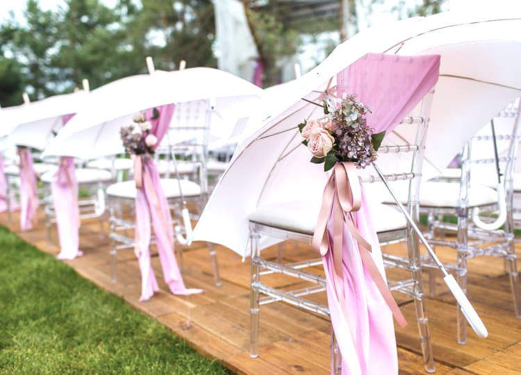 Düğün için hazırlanmış olan bahçede bulunan sandalyelerin kenarına asılmış, açık renkli şemsiyeler duruyor. Sandalyelerin kenarlarında çiçek süslemeleri ve kurdeleler bulunuyor.
