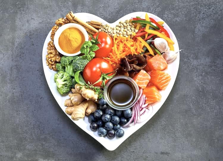 Koyu renk arka plan üzerinde kalp şeklinde bir tabak bulunuyor. Tabak içerisinde yaban mersini, brokoli, biber, balık, ceviz, domates, sarımsak ve baharat bulunuyor.