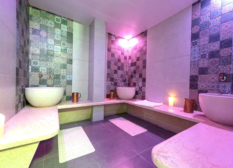 Hamam içerisinde hamam tası, mumlar bulunuyor. Hamam mor bir ışık ile aydınlatılıyor.