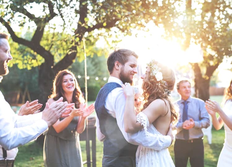 Kır düğünü yapan gelin ve damat, bahçede misafirlerin ortasında birbirlerine sarılarak dans ediyor ve gülümsüyorlar.