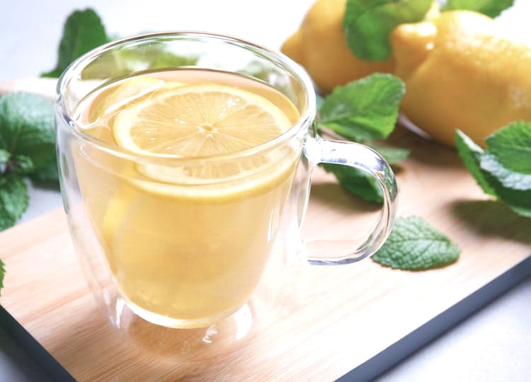 Bir bardak içerisinde su ve limon dilimleri, bardağın etrafında nane yaprakları ve limonlar bulunuyor.