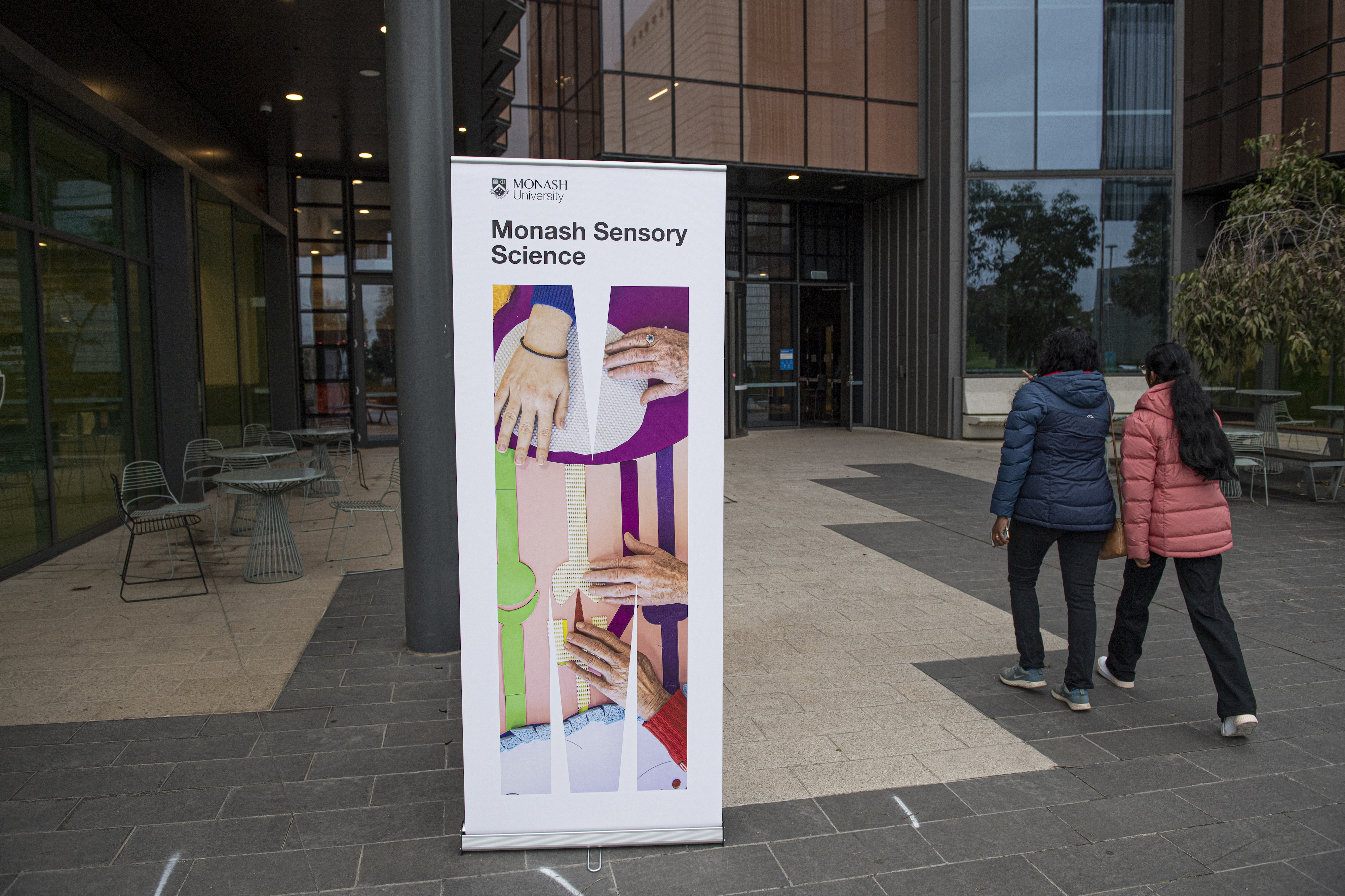 A Monash Sensory Science pull-banner in front of an entrance to a Monash University building. Two women with dark black hair wearing puffer jackets are walking towards the doorway.