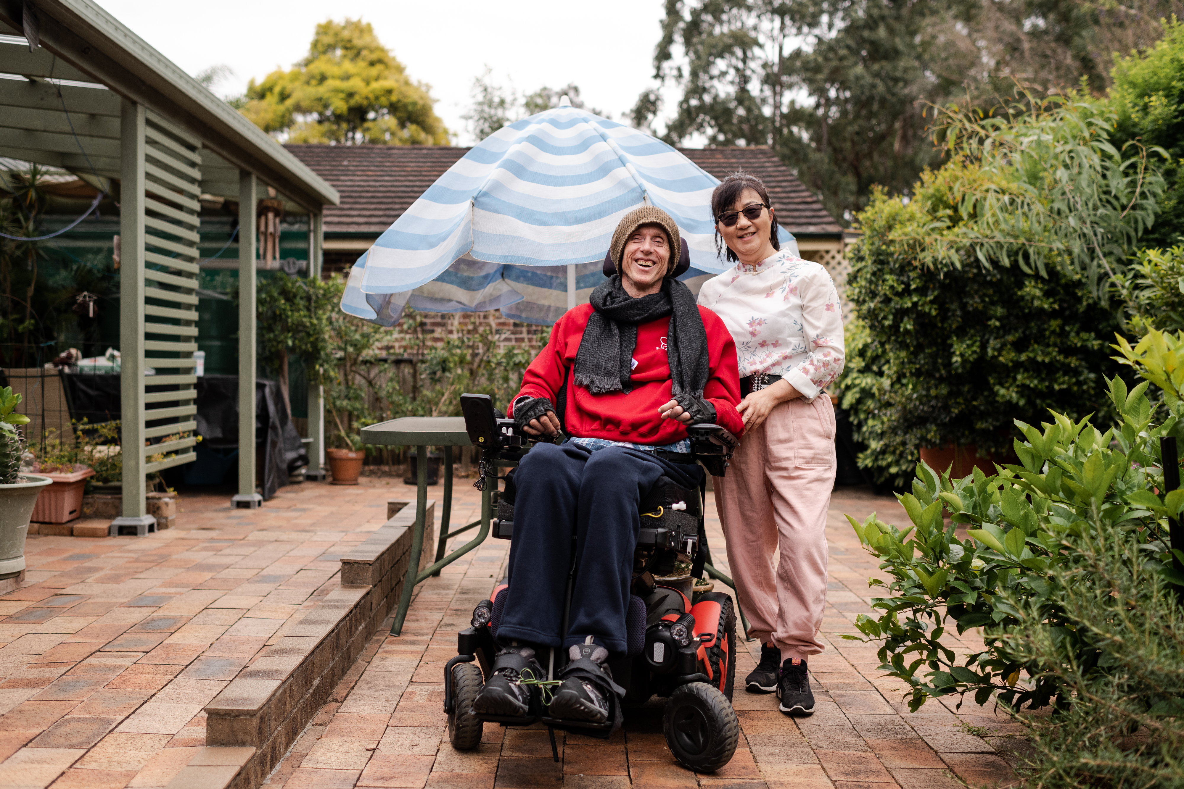 Annie and Stew are smiling together on a footpath with Stew's black labrador. Annie has short black hair, wears glasses and is wearing a white top with pink flowers and pink shorts. Stew is wearing a brown beanie, black scarf, red jumper and navy track pants. Stew is a wheelchair user.