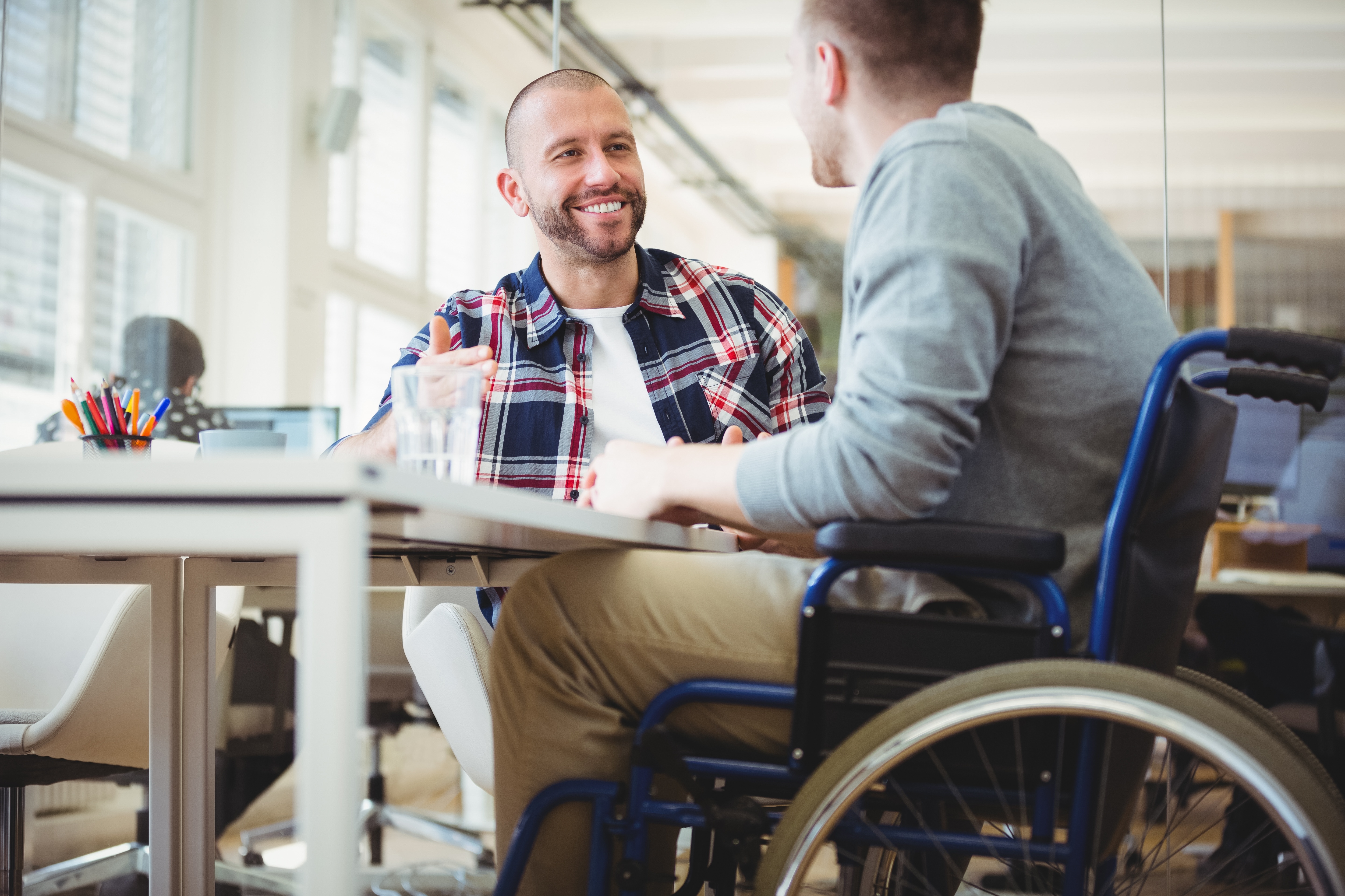 Two men with shaved short hair are sitting at a desk smiling at each other. One is wearing red and blue tartan collared shirt unbuttoned over a white t-shirt. The other is wearing a grey jumper and tan trousers and uses a wheelchair. 
