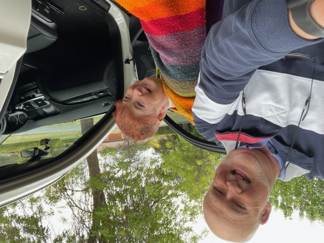 Brian and Jill are smiling, getting into a car. Brian is wearing a blue and white jumper. Jill has short red hair and is wearing a striped rainbow poncho.