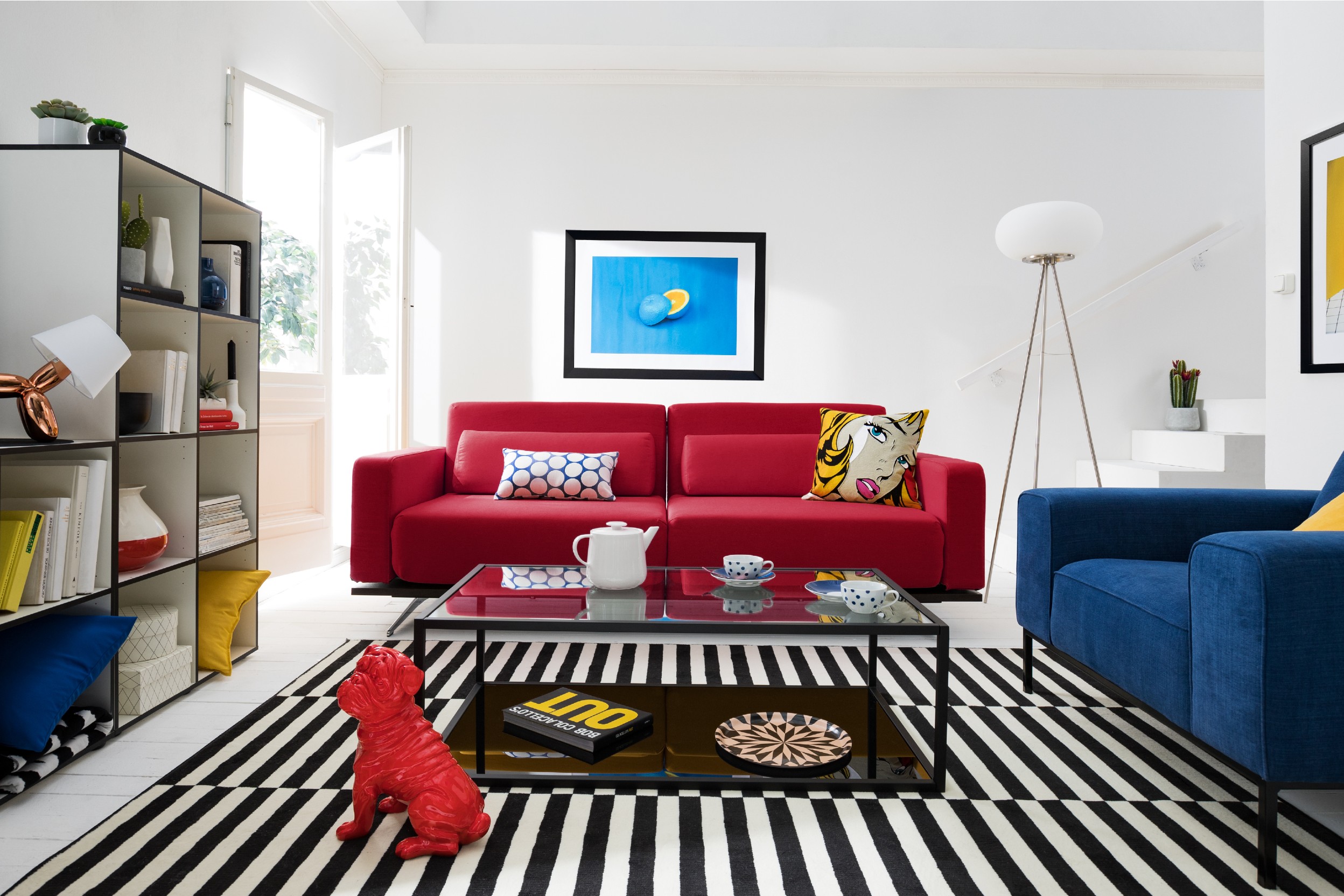 Photo d'un salon avec deux canapés, l'un rouge, l'autre bleu roi, et une table basse en verre et un tapis rayé noir et blanc
