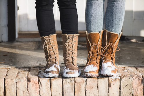 Two pairs of brown lace-up winter boots with fur lining and snow on the soles, standing side by side on a brick ledge.