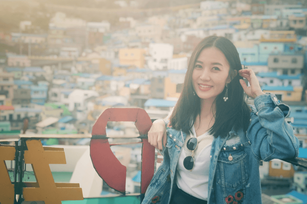 A Korean woman in a jean jacket, white shirt, and sunglasses poses in front of the buildings of Busan next to a sign in Korean.