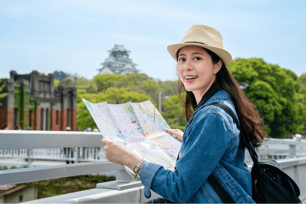 A Japanese woman wearing a sun hat looks at a map with Osaka castle in the background on a sunny day.