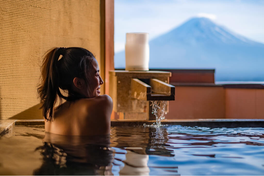 A woman smiling in a hot spring that has a view of Mt. Fuji as water pours into the hot spring