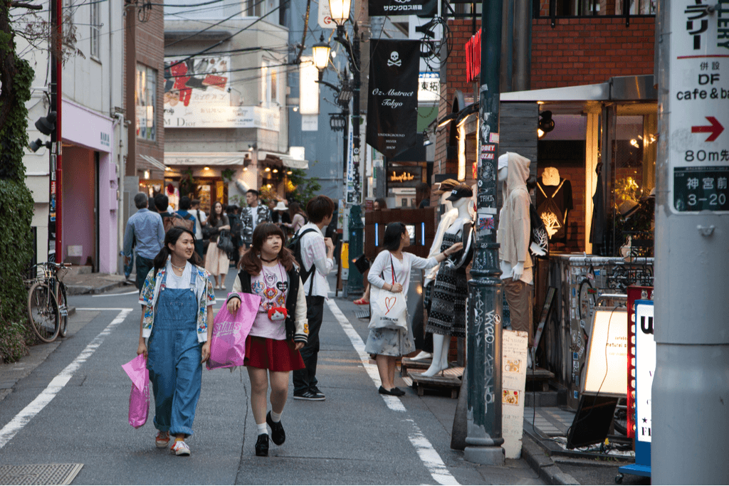 Two girls walking down the street holding shopping bags with people looking at clothes on mannequins in the background.