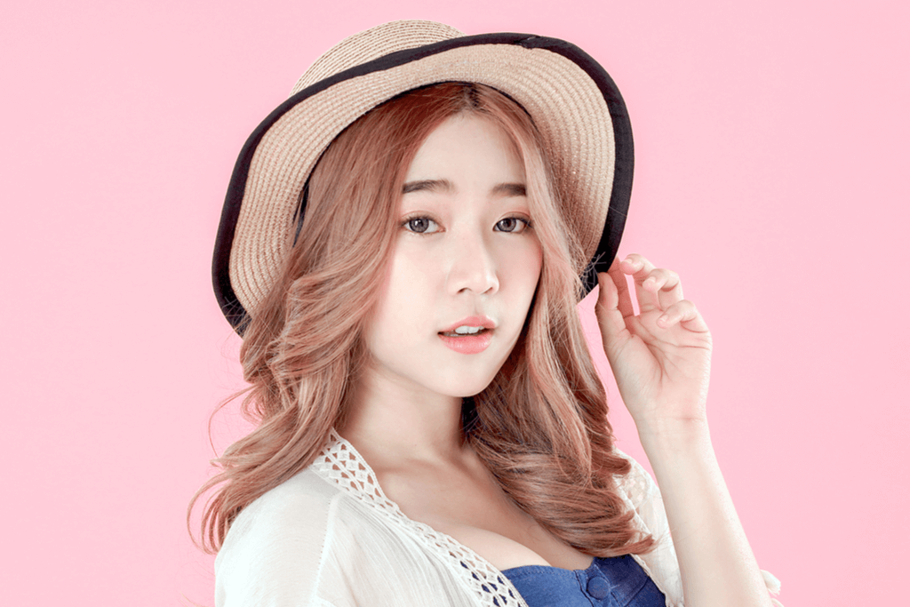 A woman in a straw hat looks at the camera as she plays with her pink hair, that is silky smooth thanks to Korean hair care products.