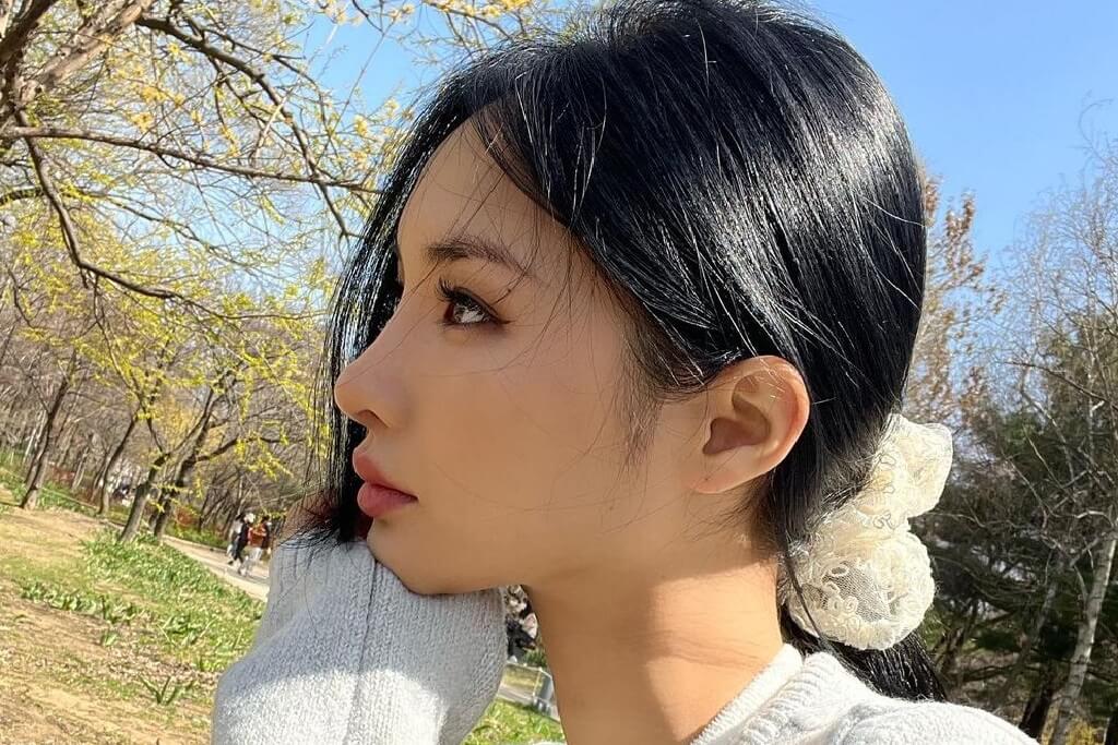 Chailee Son, a popular Korean Instagram influencer and fashion and skincare brand owner, sits in a park looking away from the camera wearinga white scrunchie and white jacket