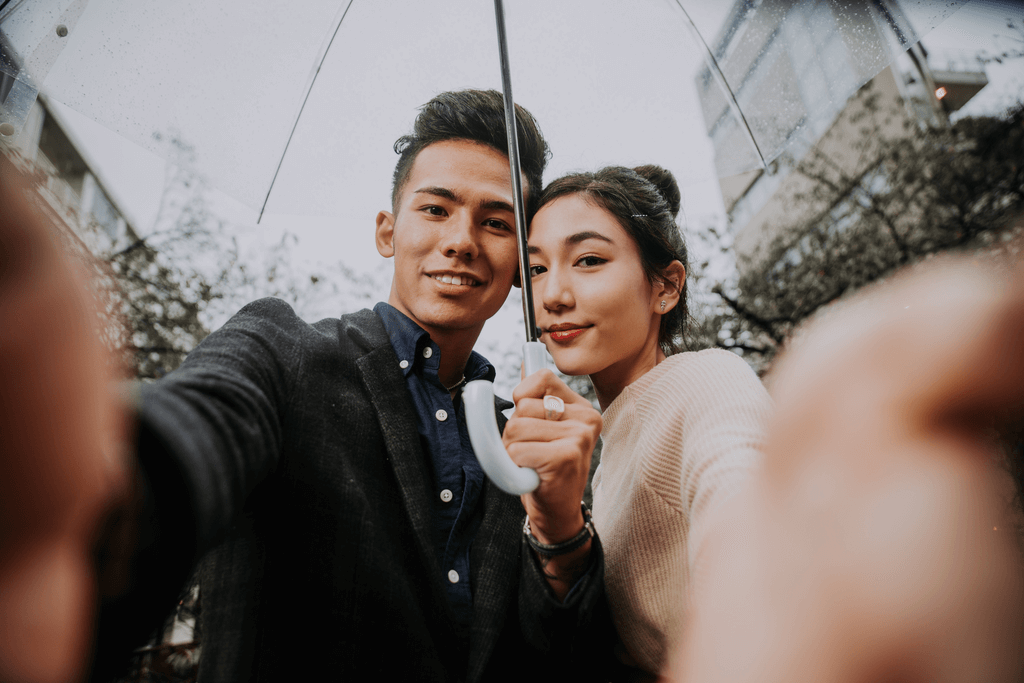 A couple holds a camera and takes a selfie together in the rain holding an umbrella