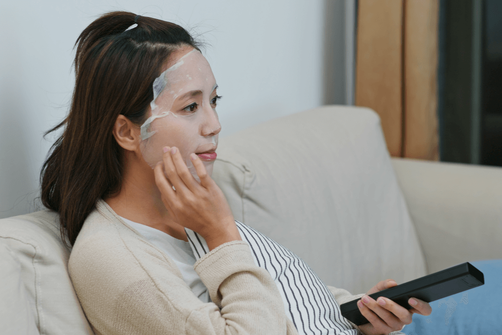 A woman flips through channels on her couch as she relaxes with a sheet mask and pats it onto her face with one hand. 