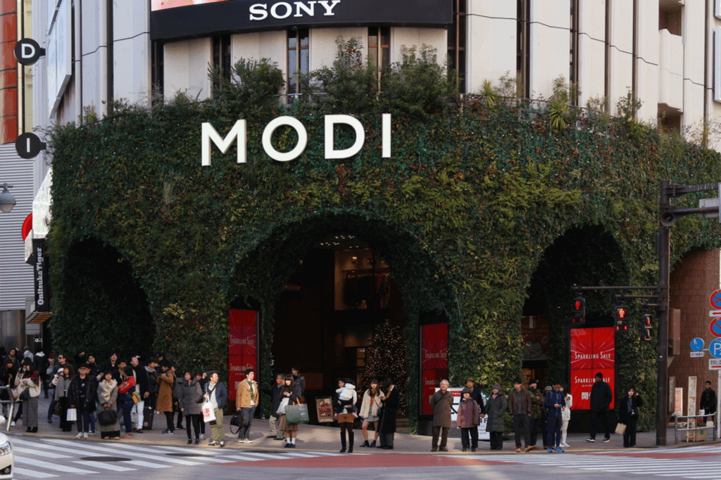 The front of the Shibuya MODI building with many leaves and greenery on the front with many people chatting and waiting for the crosswalk.