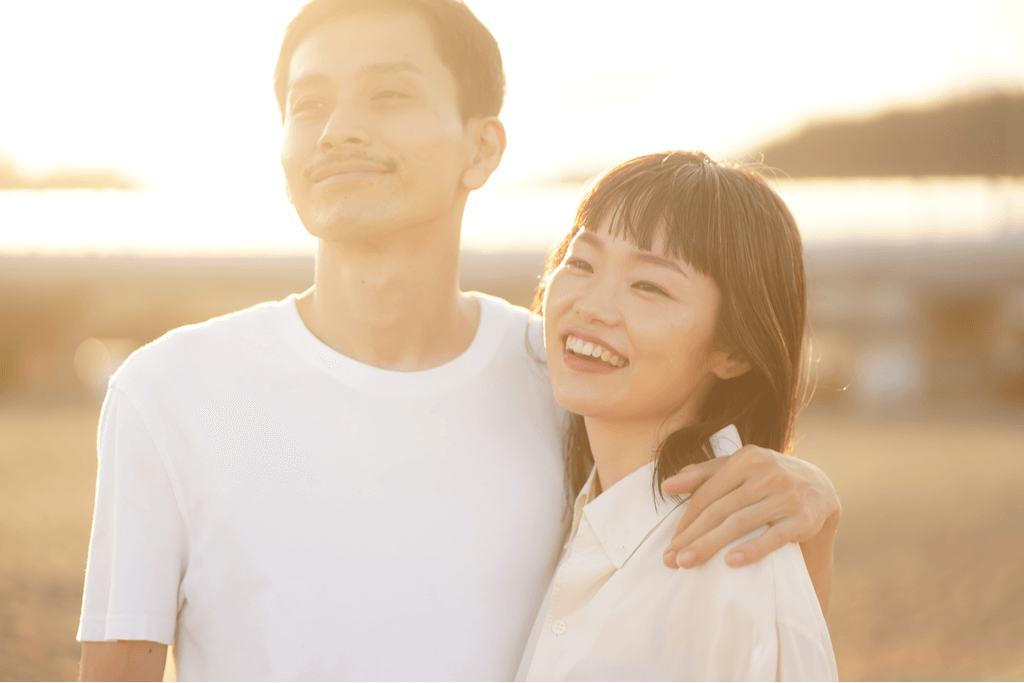 Japanese couple give a side hug on the beach with the sun starting to set in the background.