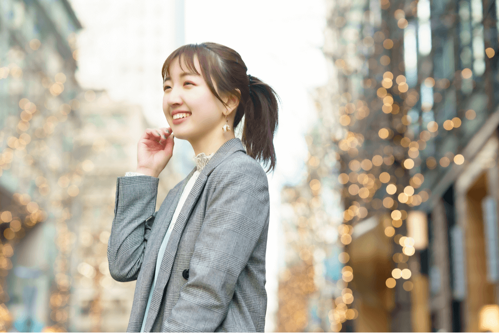 A stylish woman smiles and touches her earring as the lights from trees sit in the background on a fashion street in Tokyo