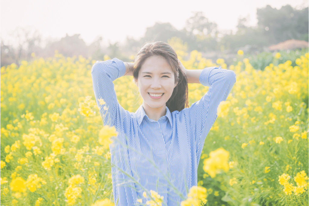 A Korean woman stands in the yellow flower fields of Jeju Island while pulling her hair back and smiling, feeling the vacation vibes.