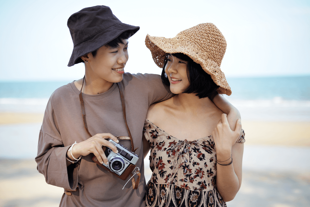 A Japanese couple walks on the beach with both women having an arm around each other while one holds a camera.