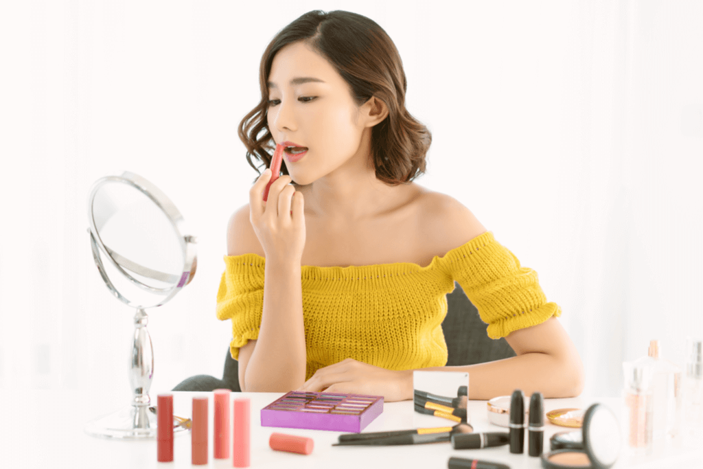 A woman applies a lip tint to her lips as she sits at her makeup station in a white room while wearing a yellow shoulderless shirt.