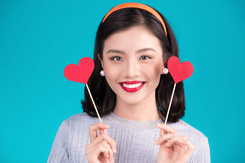 A woman stands in front of a blue background with beautiful Japanese makeup holding two hearts.