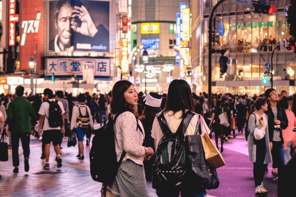 Two women stand at Shibuya Crossing at night with many different people in the background chatting and walking around with many bright buildings behind them.