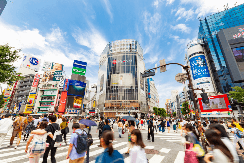 Many people walk through the Shibuya crossing with many buildings in the background and blue skies overhead.