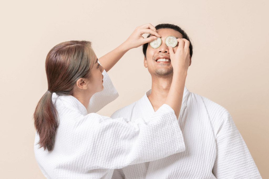 A woman cutely places cucumbers over a man's eyes as they both wear robes in front of a beige background.