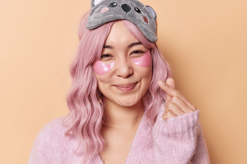 A young woman in pink pajamas and a Koala eye mask makes a heart mark with her fingers as she smiles while having under eye patches on.