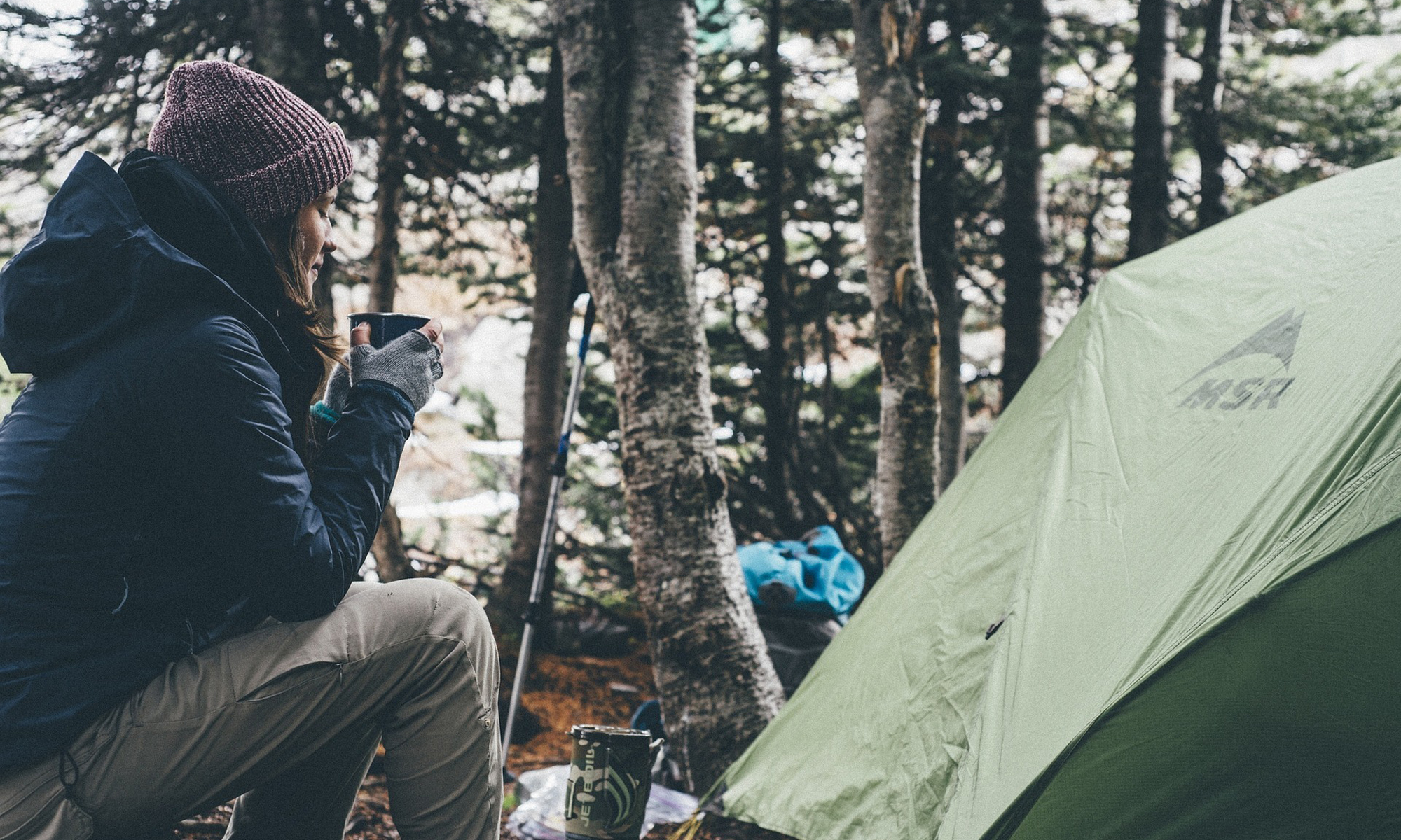 Woman camping in the woods and drinking coffee in the morning by her tent.