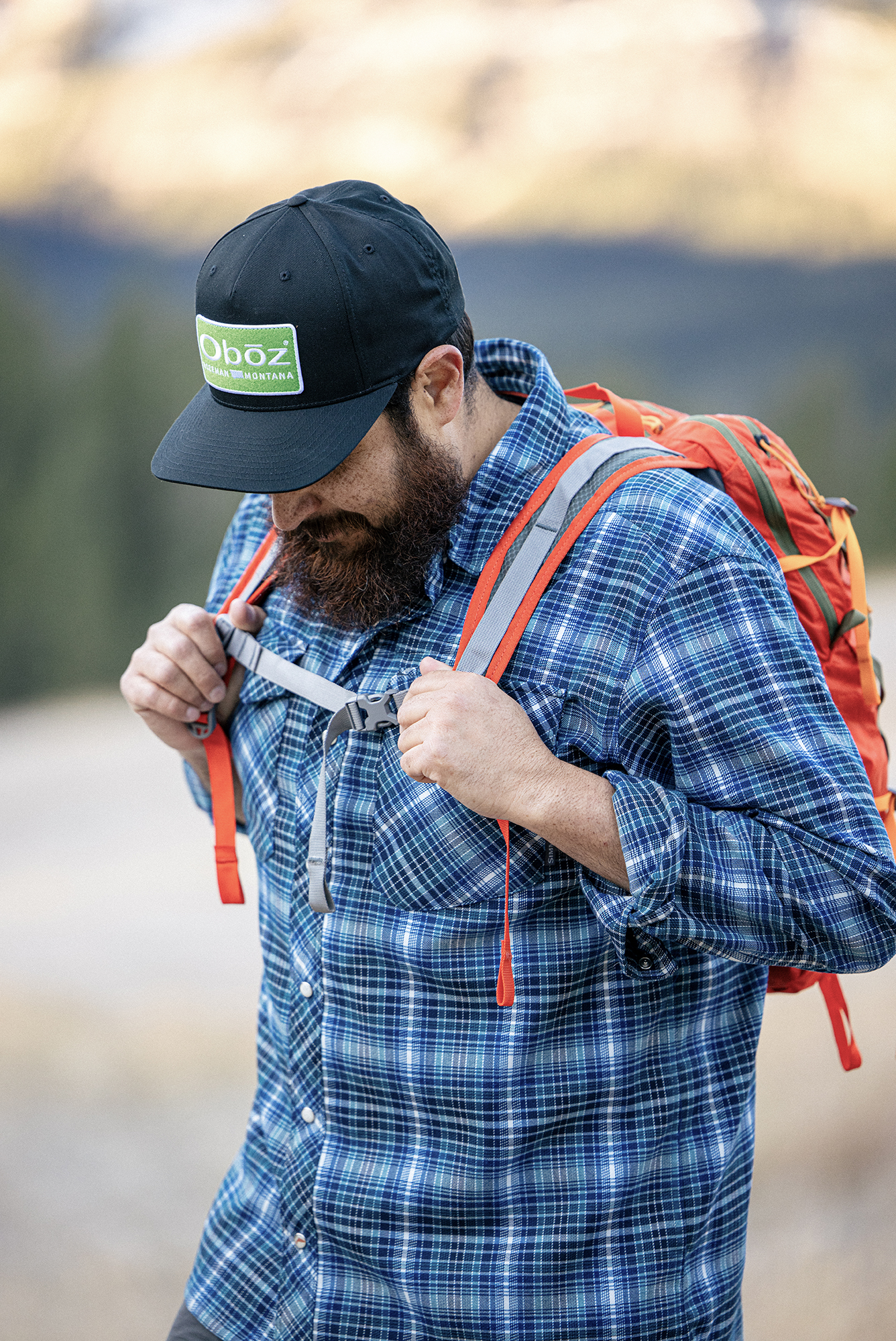 Man adjusting his backpack chest strap while hiking in an Oboz Footwear hat.