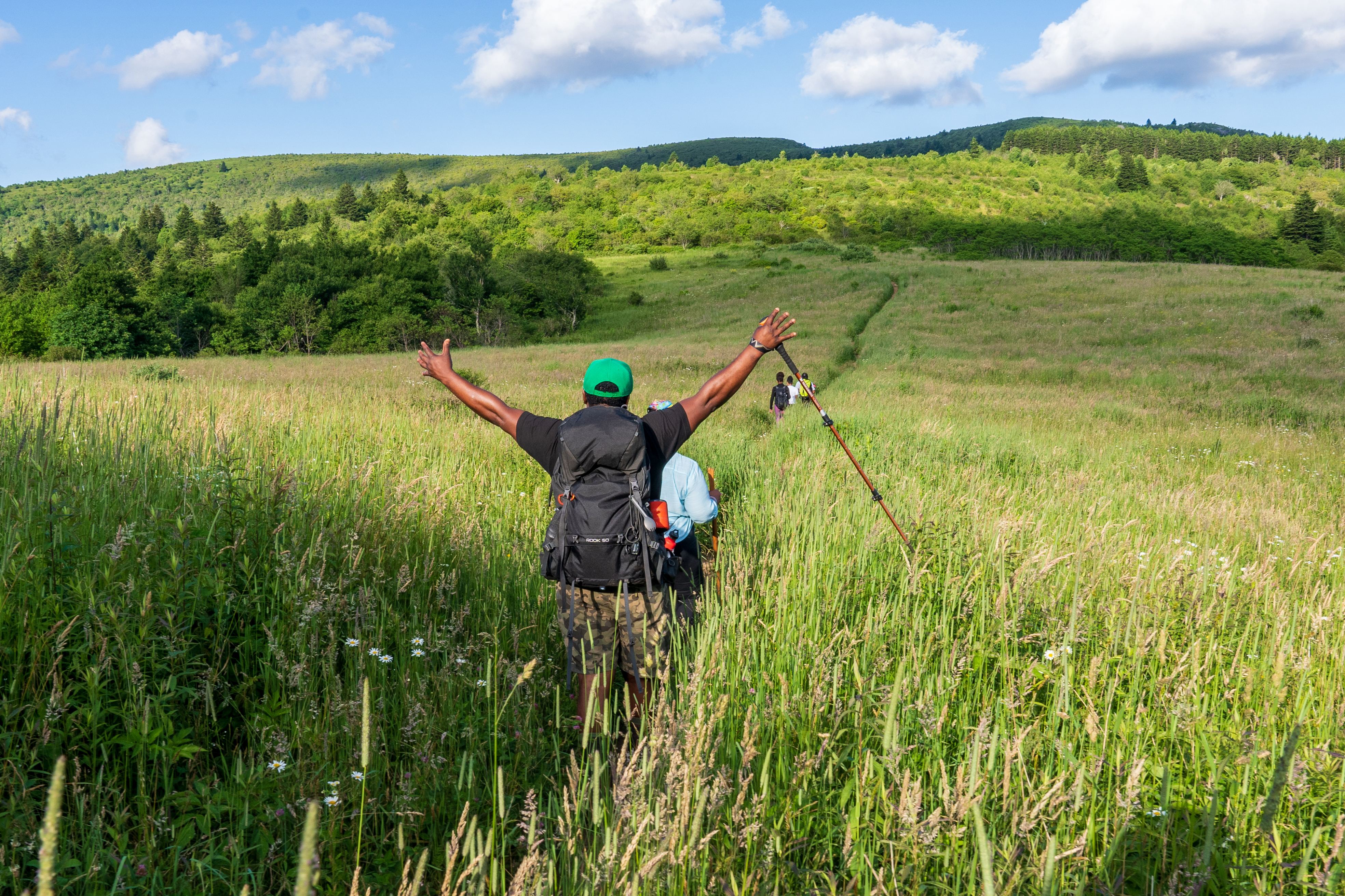Hiker is raising hands victoriously while walking through a grassy plain.