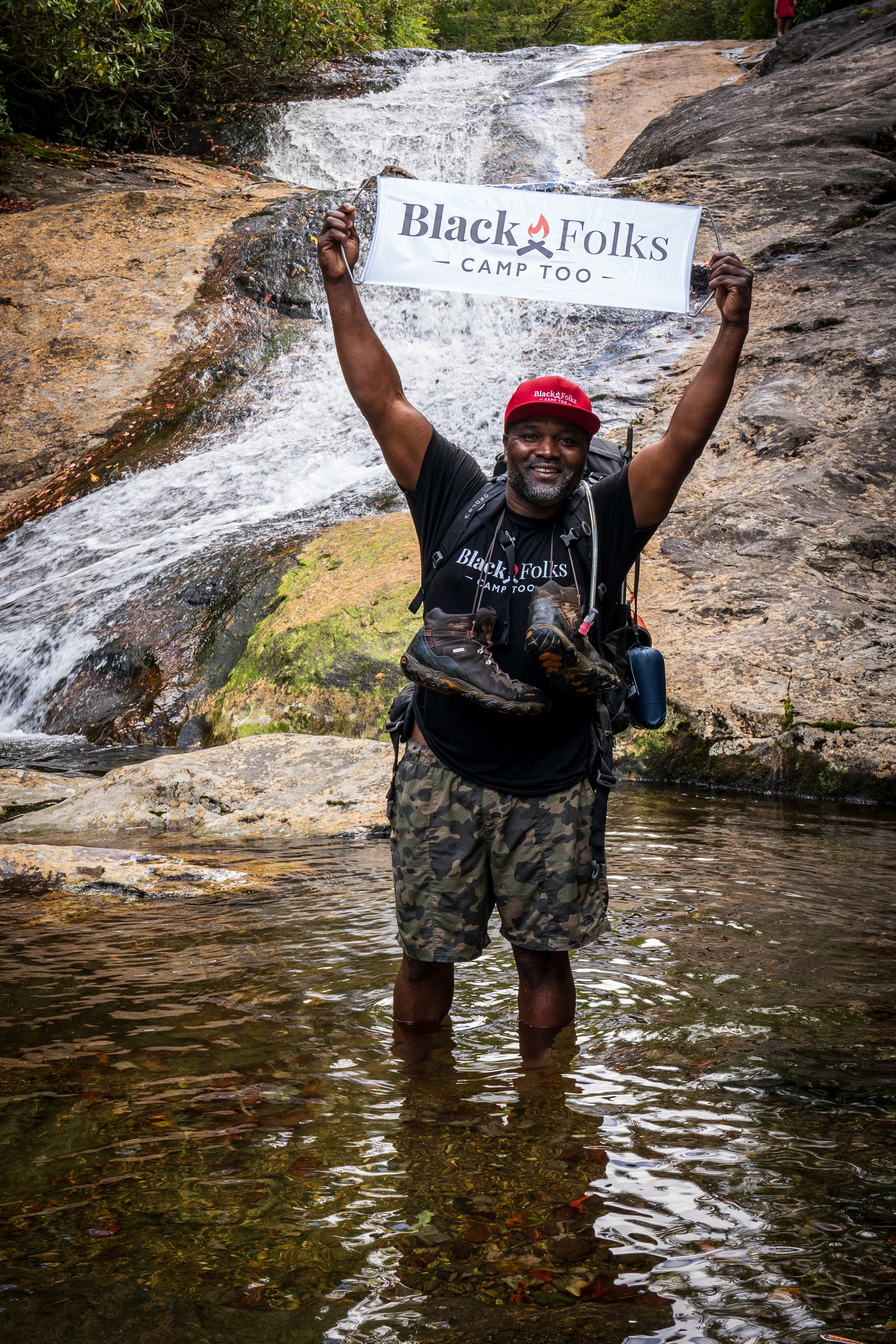 Earl Hunter in front of a waterfall holding up a Black Folks Camp Too sign