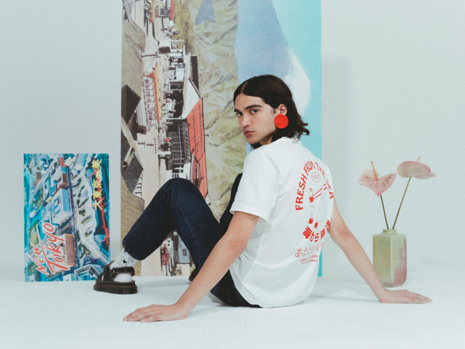 A man wearing a white T-shirt with Japanese characters and jeans, sitting on the floor.
