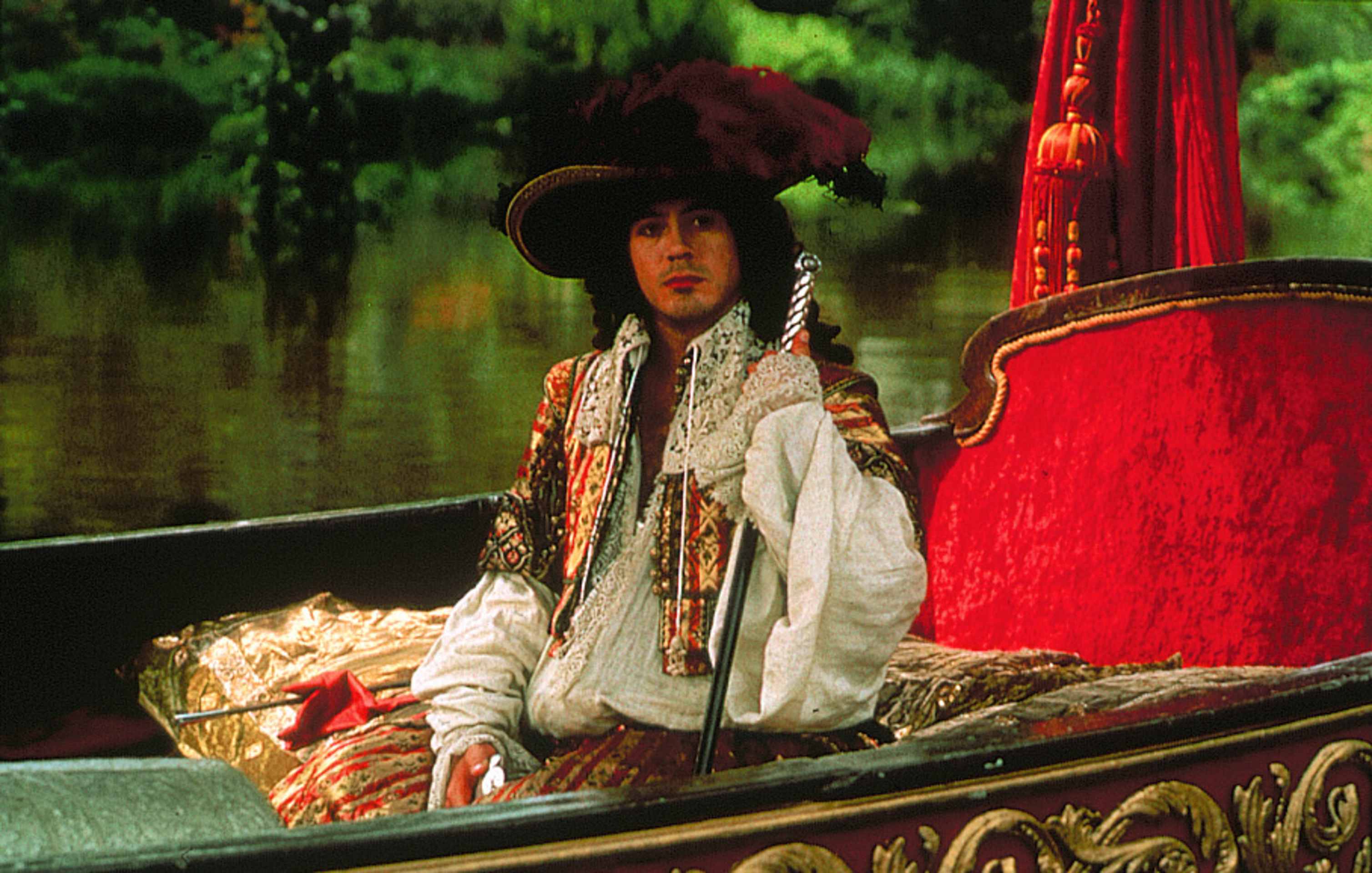 A white man in 17th Century regal, flamboyant attire, sitting in a plush lake boat