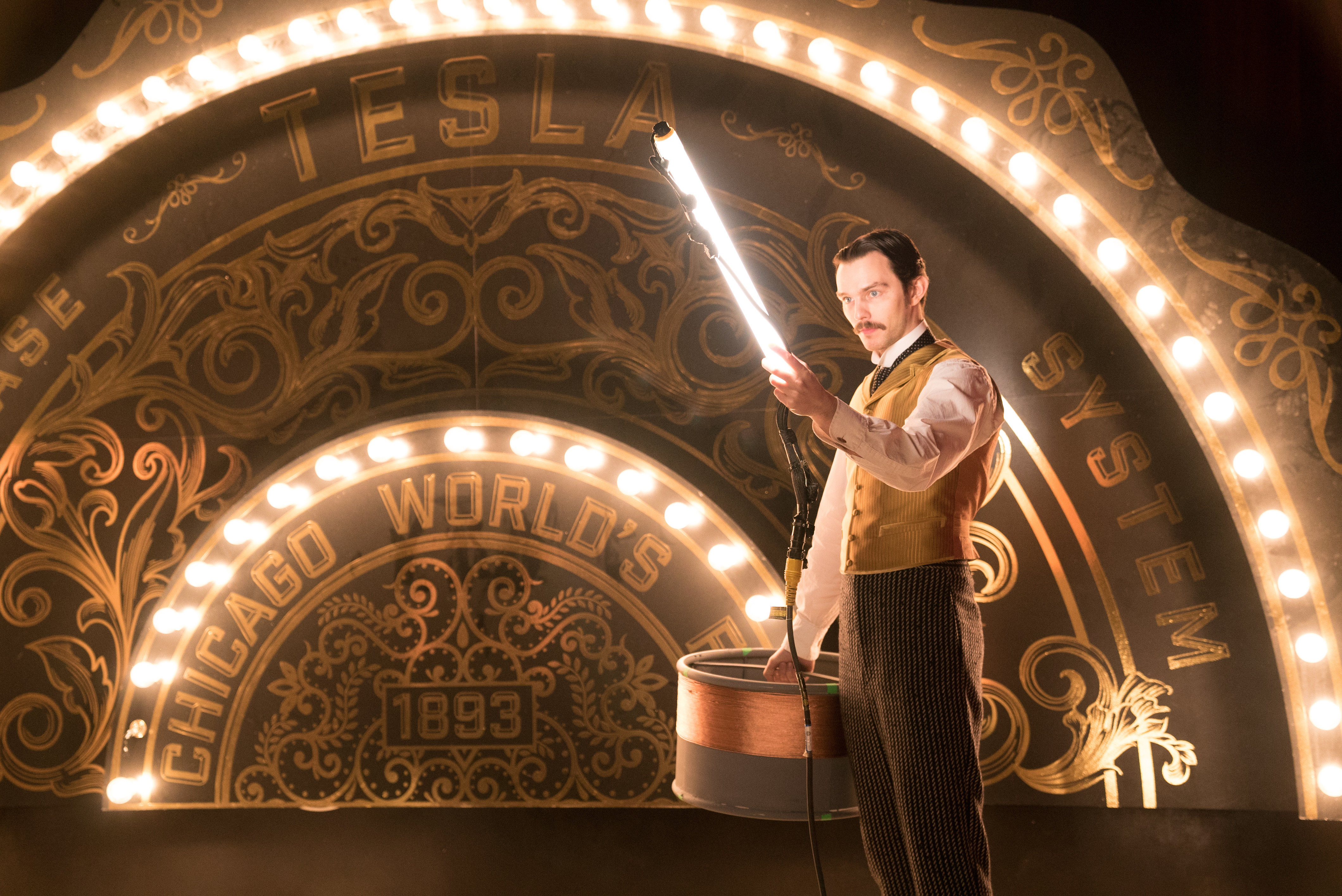 A young white man wearing a smart 1880's suit, holding a tube of light, with an arch of light bulbs over a sign reading 'TESLA'